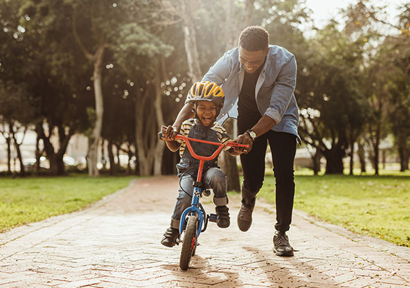 A dad running and pushing his son on a bicycle. He is teaching his son to ride without training wheels. 