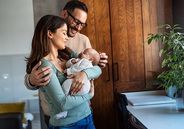 A mom holding a baby in her arms while dad smiles and looks at the baby.
